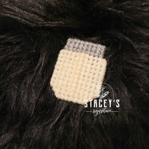 May include: A small crocheted jar with a white body and a gray lid. The jar is on a black fuzzy surface. The text "STACEY'S signature" is below the jar.