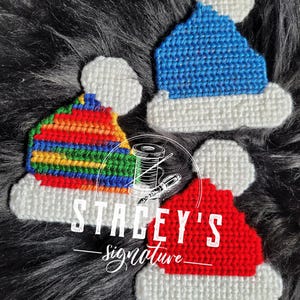 May include: Three handmade Christmas hat ornaments in blue, red, and rainbow colors with white trim and pom-poms. The ornaments are displayed on a dark, textured background. The text "STACEY'S signature" is visible.