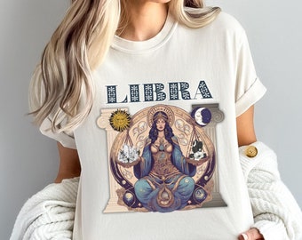 May include: A white t-shirt with a graphic of a woman sitting on a throne with a scale in her hands. The graphic is surrounded by stars, moons, and the word "LIBRA" in blue lettering.