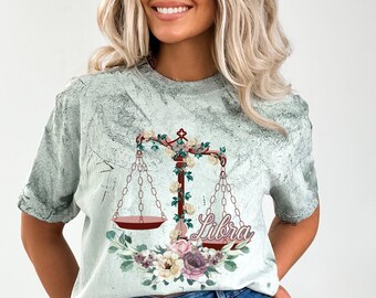 Può includere: Una maglietta verde chiaro con un aspetto usurato che presenta un design floreale con una bilancia e la parola "Libra" in corsivo.