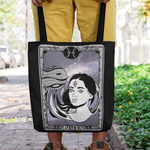 Könnte beinhalten: Schwarze Tragetasche mit einer weißen und grauen Illustration einer Frau mit einem dritten Auge und einem Planeten im Hintergrund. Die Illustration ist von einem weißen Rahmen umgeben und das Wort "Pisces" ist unter der Illustration gedruckt.