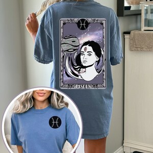 Könnte beinhalten: Ein blaues T-Shirt mit einem Grafikdruck einer Tarotkarte, die eine Frau mit einem dritten Auge zeigt und das Wort "Pisces" auf der Rückseite. Die Vorderseite des Shirts hat ein kleines schwarzes Fische-Symbol.