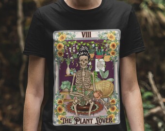 Puede incluir: Camiseta negra con un gráfico de un esqueleto sentado en una silla rodeado de plantas. El gráfico está en el estilo de una carta de tarot y dice "VIII The Plant Lover".
