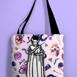 May include: A white tote bag with black straps features a black and white illustration of a Frankenstein's monster on a pink background with a repeating pattern of skulls, snakes, flowers, and other gothic imagery.