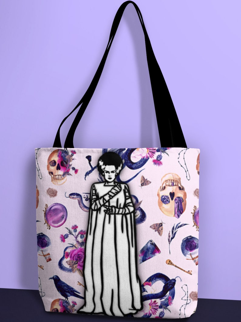 May include: A white tote bag with black straps features a black and white illustration of a Frankenstein's monster on a pink background with a repeating pattern of skulls, snakes, flowers, and other gothic imagery.