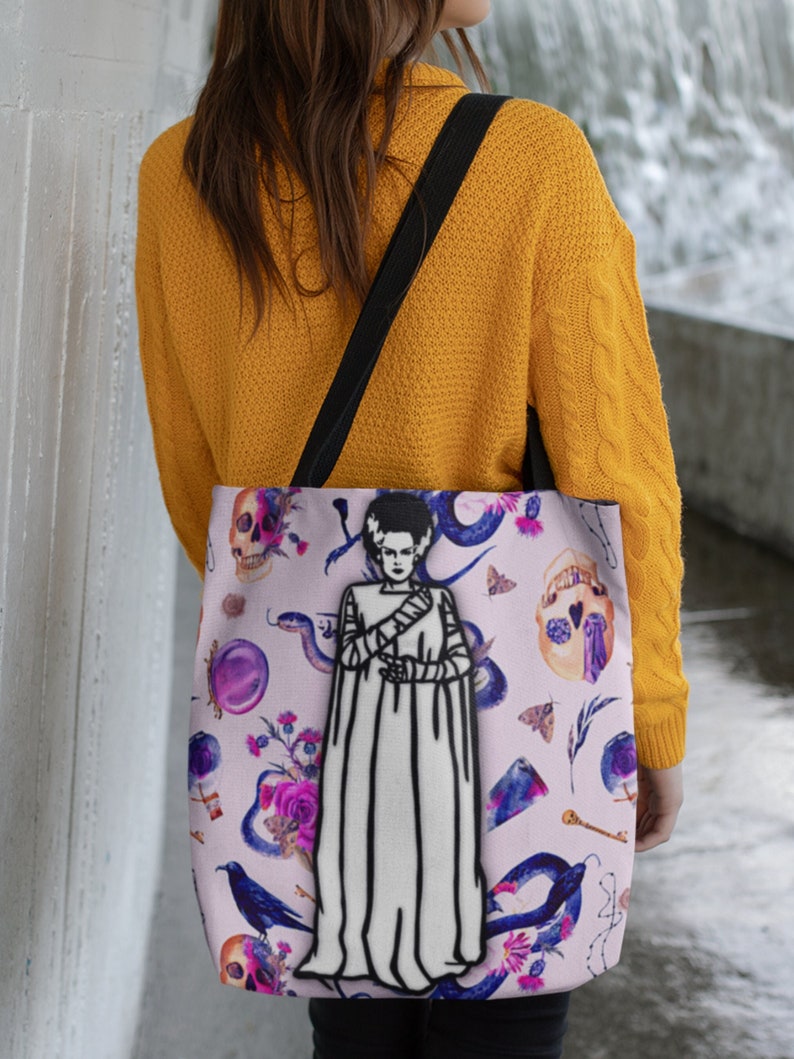 May include: A pink tote bag with a black strap features a white illustration of a woman with a white dress and black hair. The background is a pink floral pattern with snakes, skulls, and other gothic elements.