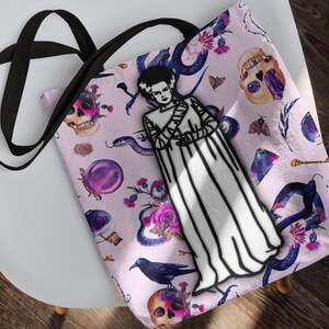 May include: A pink tote bag with a black strap features a whimsical design of a black and white illustration of a monster in a white gown, surrounded by snakes, skulls, flowers, and other gothic elements.
