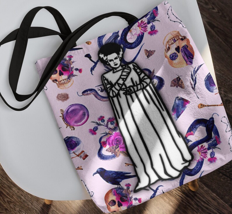 May include: A pink tote bag with a black strap features a whimsical design of a black and white illustration of a monster in a white gown, surrounded by snakes, skulls, flowers, and other gothic elements.