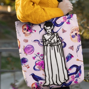 May include: A pink tote bag with a black and white illustration of Frankenstein's monster. The bag is decorated with a variety of other spooky and whimsical illustrations, including skulls, snakes, flowers, and a raven.