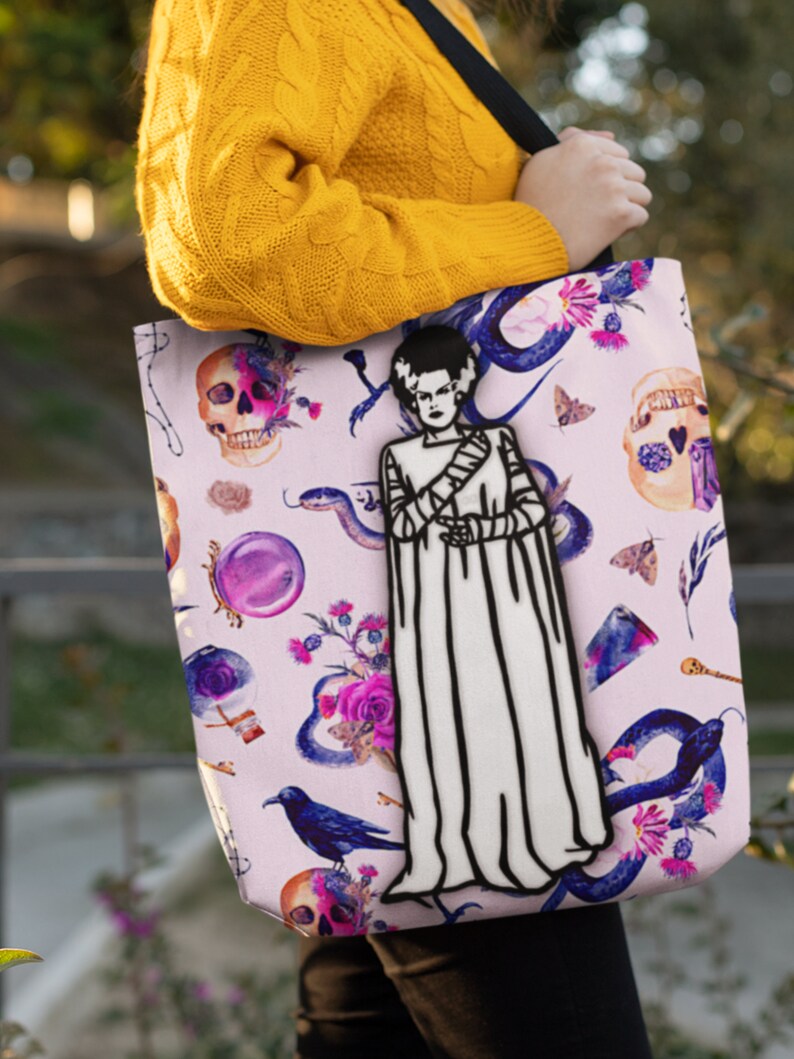 May include: A pink tote bag with a black and white illustration of Frankenstein's monster. The bag is decorated with a variety of other spooky and whimsical illustrations, including skulls, snakes, flowers, and a raven.