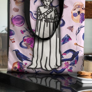 May include: A pink tote bag with a black and white illustration of a monster bride. The bag is decorated with a floral pattern, skulls, and snakes.
