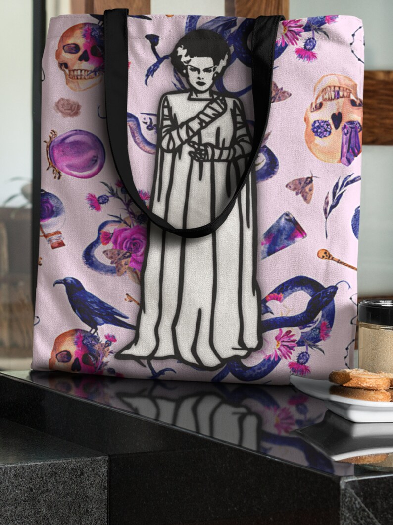 May include: A pink tote bag with a black and white illustration of a monster bride. The bag is decorated with a floral pattern, skulls, and snakes.