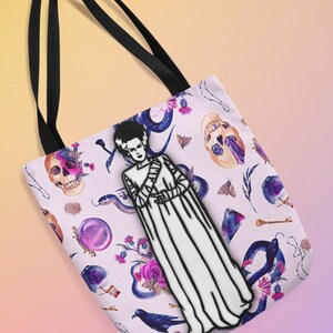 May include: A white tote bag with black straps features a black and white illustration of a monster with a white gown and a floral, skull, and snake pattern.