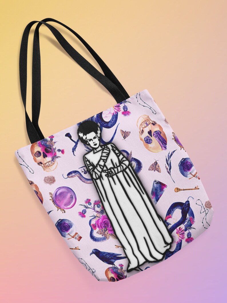 May include: A white tote bag with black straps features a black and white illustration of a monster with a white gown and a floral, skull, and snake pattern.