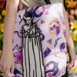 May include: A pink tote bag with a black and white illustration of a woman with a monster-like appearance. The bag has a floral and skull design with purple, blue, and green accents.