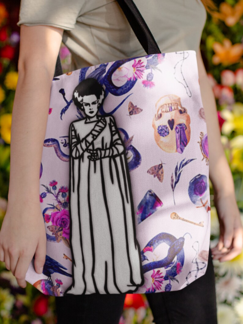 May include: A pink tote bag with a black and white illustration of a woman with a monster-like appearance. The bag has a floral and skull design with purple, blue, and green accents.