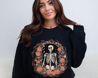May include: Black sweatshirt featuring a graphic print of a skeleton sitting within a wreath of orange flowers. The skeleton is wearing a skull.