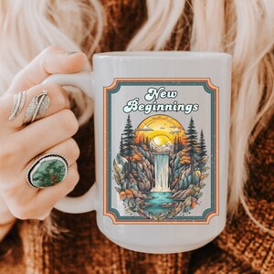 Könnte beinhalten: Weiße Keramiktasse mit einer bunten Illustration einer Wasserfall- und Waldszene. Die Tasse trägt den Text "New Beginnings" in einer Retro-Schrift.