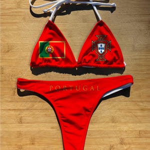 May include: Red bikini set featuring the Portuguese flag and coat of arms. The top has triangle cups with white straps. The bottom has the word "PORTUGAL" printed across the front. The bikini is displayed on a wooden surface.