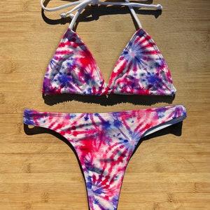 May include: A white bikini with a tie-dye pattern in shades of pink, blue, and white. The bikini top is a triangle style with white straps. The bikini bottom is a thong style.