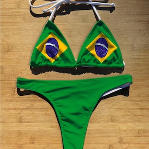May include: A green bikini set featuring the Brazilian flag design. The top has triangle cups with the flag's emblem, and the bottoms are a solid green thong style. White string ties complete the top.