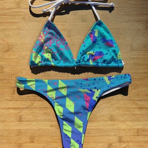 Può includere: Un bikini blu e verde con un motivo geometrico. La parte superiore è di stile triangolare con lacci bianchi. La parte inferiore è di stile a taglio alto con un retro a tanga.