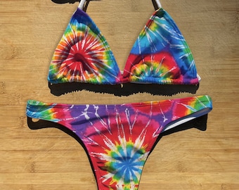 May include: A tie-dye bikini set featuring a halter top and a thong-style bottom. The colorful pattern includes red, orange, yellow, green, blue, and purple hues. The top has white straps, and the set is displayed on a wooden surface.