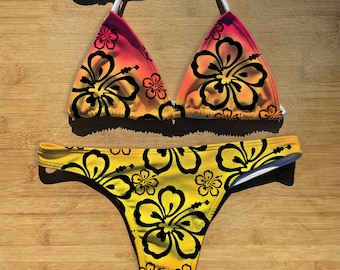May include: A yellow and pink bikini with a black hibiscus flower print. The bikini top is a triangle style with adjustable straps. The bikini bottom is a cheeky style with a high cut leg.