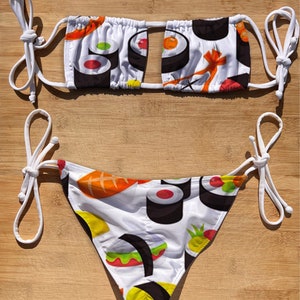 以下が含まれることがあります： 寿司柄の白いビキニ。トップはバンダナスタイルで、フロントにカットアウトがあります。ボトムはチークスタイルで、調節可能な紐が付いています。