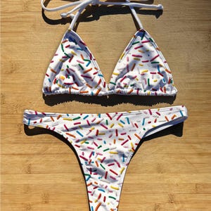Può includere: Bikini bianco con motivo a spruzzi colorati. La parte superiore è a triangolo con lacci bianchi, e la parte inferiore è a perizoma. Il bikini è esposto su una superficie di legno.