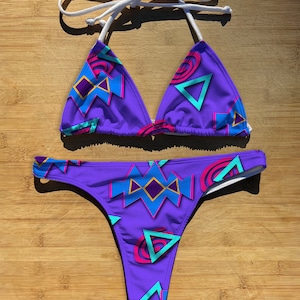 May include: Purple bikini with a geometric pattern in shades of blue, pink, and yellow. The bikini top is a triangle style with white ties. The bikini bottom is a high-cut style.