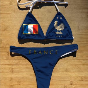 May include: A blue bikini set featuring the French flag and a golden rooster emblem. The top has white straps and the bottom has the word "FRANCE" in gold lettering. The swimsuit is displayed on a wooden surface.