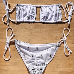 Op de afbeelding: Witte bikini set met een zwart paisley patroon. De top heeft een sleutelgat ontwerp en verstelbare bandjes. De broek is een klassieke snit met strikzijden. De stof is gerimpeld.