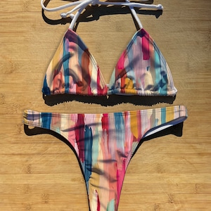 May include: A colorful bikini with a triangle top and a high-cut bottom. The fabric features a watercolor-like pattern with abstract brushstrokes in shades of pink, blue, yellow, and orange.