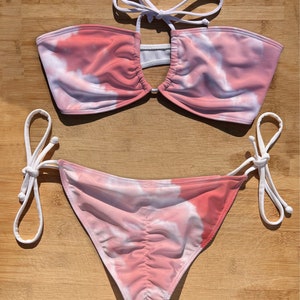 Puede incluir: Un top y una parte inferior de bikini de color rosa y blanco con estampado tie-dye y tirantes blancos. El top tiene un escote de ojo de cerradura en la parte delantera y la parte inferior tiene lazos ajustables a los lados.