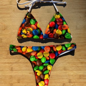 Può includere: Un bikini nero con una stampa colorata di M&M's. La parte superiore è di stile triangolare con spalline regolabili. La parte inferiore è di stile audace con vita bassa.