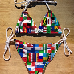 May include: A two-piece bikini set with a vibrant patchwork design featuring various national flags. The top has triangle cups with white tie straps, and the bottoms have a classic cut with matching tie sides. The swimwear is set against a wooden background.