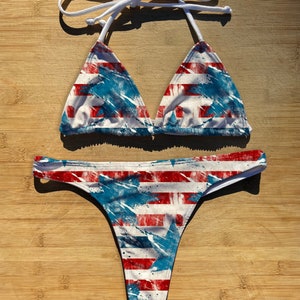 May include: A red, white, and blue bikini with a distressed American flag print. The top is a triangle style with adjustable straps. The bottom is a cheeky style with a high cut leg.