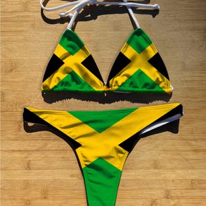 May include: A two-piece bikini set featuring the Jamaican flag design. The top is a triangle style with white straps, and the bottom is a thong cut. The colors include green, yellow, and black.