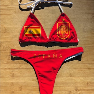 May include: Red bikini set featuring the Spanish flag and coat of arms. The triangle top has white straps and the bottom has the word "España" in gold lettering. A patriotic swimwear option.