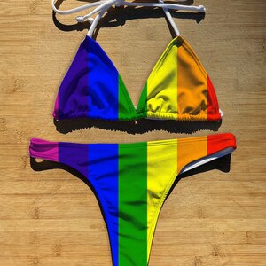 May include: A rainbow striped bikini top and bottom. The top is a triangle style with white ties. The bottom is a cheeky style with a low rise.