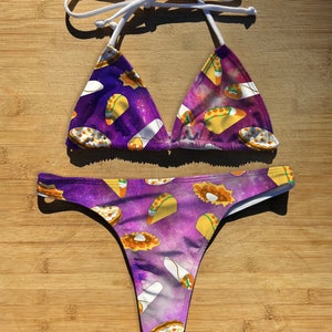 May include: A two-piece bikini set featuring a purple galaxy print with tacos and burritos. The triangle top has white straps, and the bottoms are a thong style. The swimwear is designed for a fun, food-themed look.