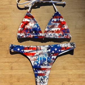 May include: A two-piece bikini set featuring a patriotic design. The top is a triangle style with white ties, and the bottoms are a thong cut. The fabric is printed with a red, white, and blue abstract pattern with stars and stripes.