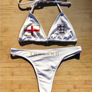 May include: White bikini set featuring the England flag and crest. The top has triangle cups with the flag and crest, and the bottoms have the word "ENGLAND" in gold lettering. The bikini is displayed on a wooden surface.