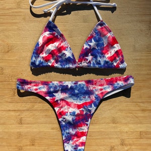 May include: A red, white, and blue bikini with a tie-dye pattern featuring stars. The bikini top is a triangle style with adjustable straps. The bikini bottom is a thong style.