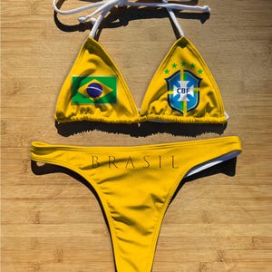 May include: Yellow bikini set featuring the Brazilian flag and CBF logo on the top. The bottom of the bikini has the word "BRASIL" printed across the front. The bikini has white straps and is displayed on a wooden surface.