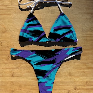 Può includere: Un bikini blu, nero e viola con un motivo a pennellate. La parte superiore del bikini è di stile triangolare con lacci bianchi. La parte inferiore del bikini è di stile a taglio alto.