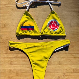 May include: Yellow bikini set with white string ties. The triangle top features the Colombian flag and a soccer ball emblem. The bottom has the word "COLOMBIA" printed across the front. The swimsuit is displayed on a wooden surface.