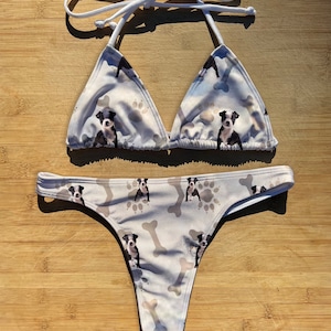 Può includere: Un bikini bianco con un motivo ripetuto di cuccioli, impronte di zampe e ossa. Il top è a fascia e la parte inferiore è a perizoma. Il bikini è realizzato in un tessuto morbido e leggero.