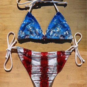 May include: A white bikini with red and blue tie-dye patterns. The top is a triangle style with white straps. The bottom has a high-cut leg and a tie-side design.